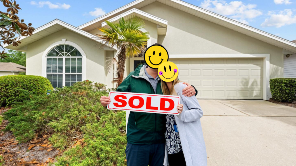 A joyful couple with smiley face emojis covering their faces stands in front of a recently purchased one-story home in Gainesville, FL, holding a bold "SOLD" sign. The home features a white garage door, arched windows, and a tropical palm tree to the side.