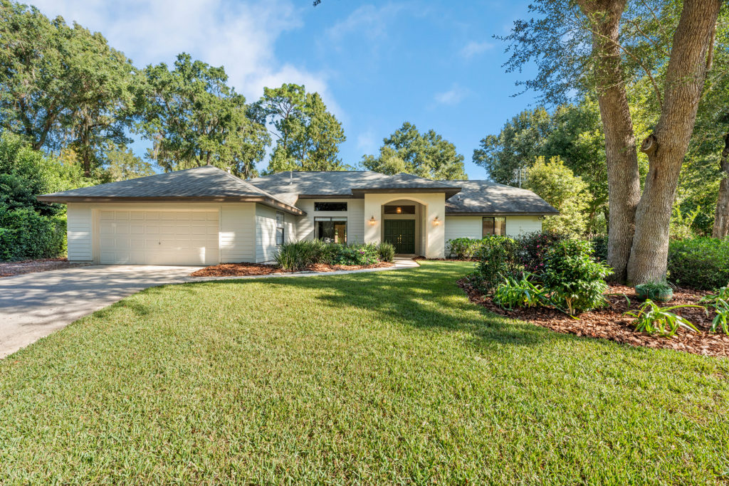 One-story home with a gray facade, attached garage, and well-maintained lawn surrounded by mature trees in Gainesville FL.